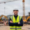 Ingeniero con casco y chaleco en sitio de construcción con maquinaria pesada y grúas en el fondo.