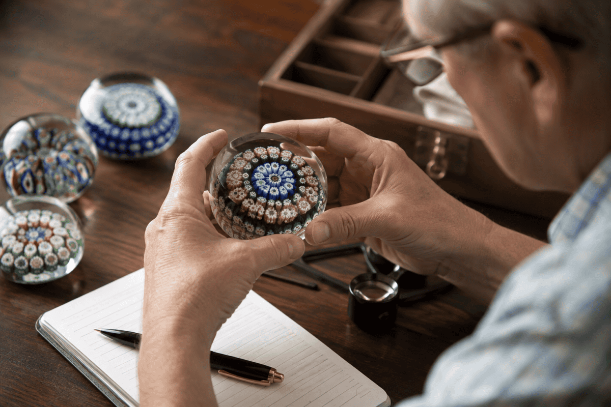 Collector carefully examining a Scottish millefiori glass paperweight beside other studio glass examples using natural light to assess craftsmanship and condition.
