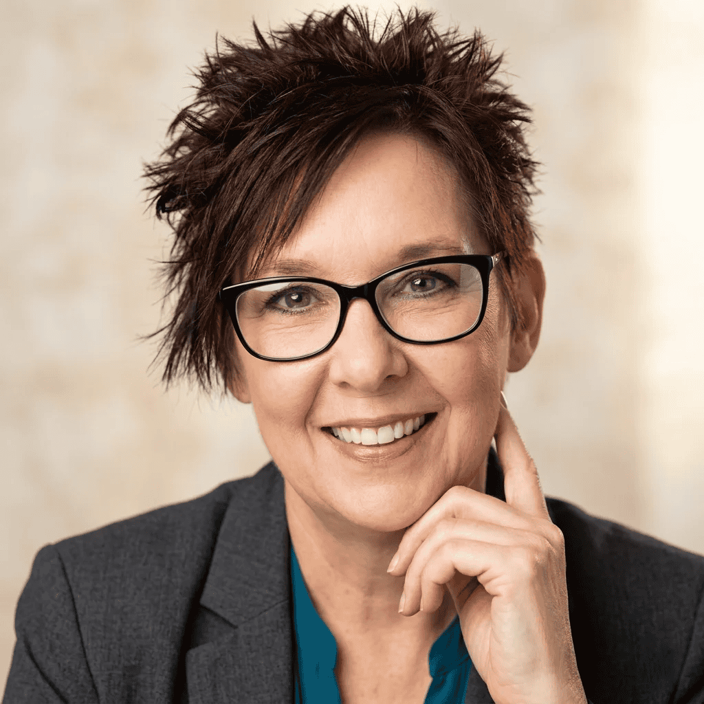 Smiling woman with short spiky hair, glasses, and a gray blazer poses with hand to chin.