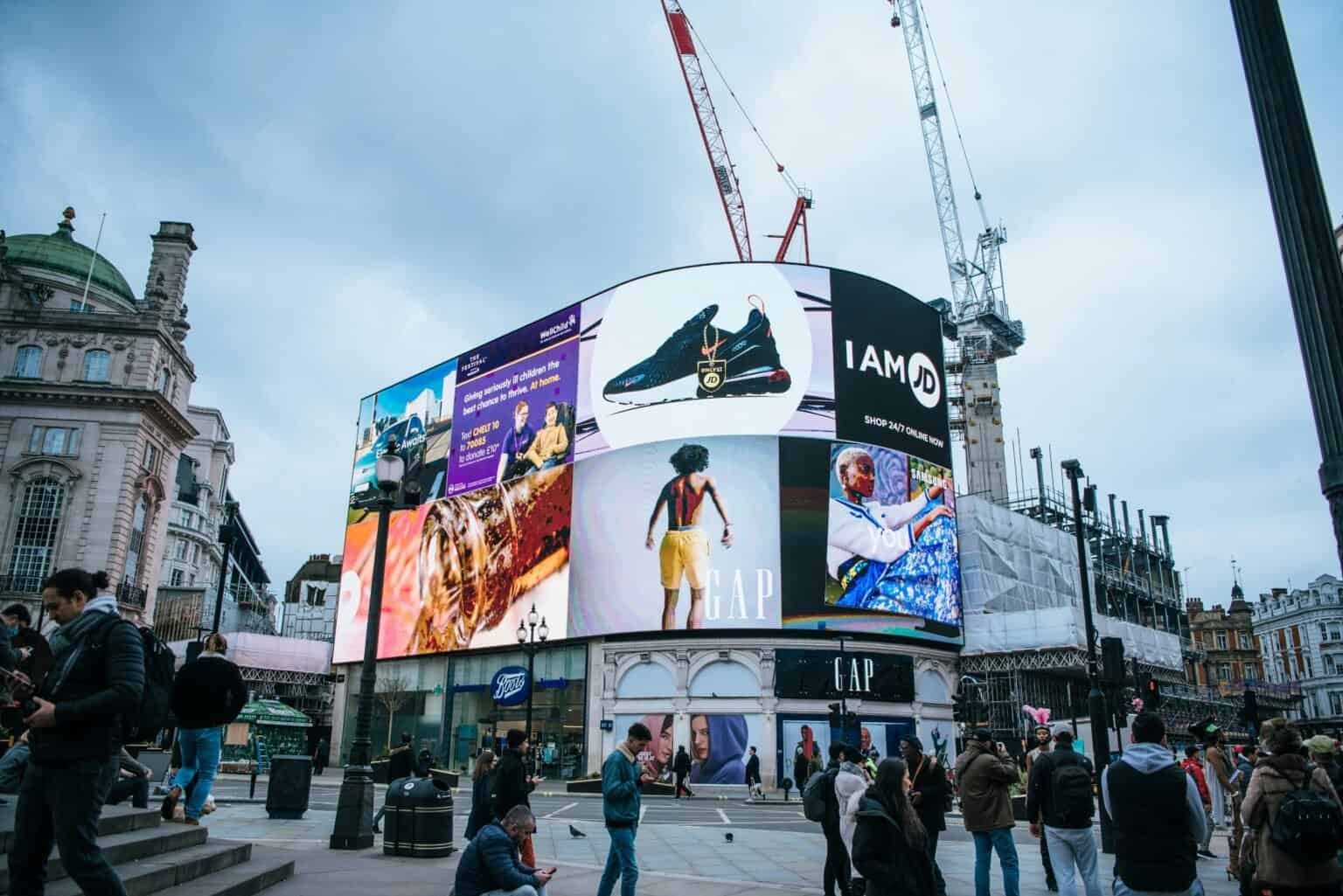Piccadilly Circus, London, with large digital billboards displaying ads for JD Sports, Boots, and Gap.