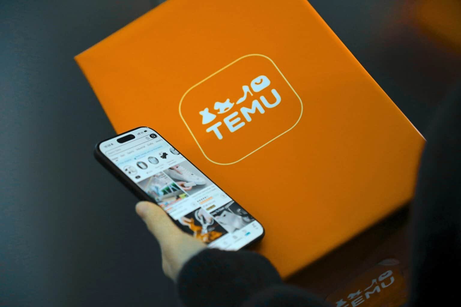 Person holding a smartphone displaying the Temu app next to a Temu box.