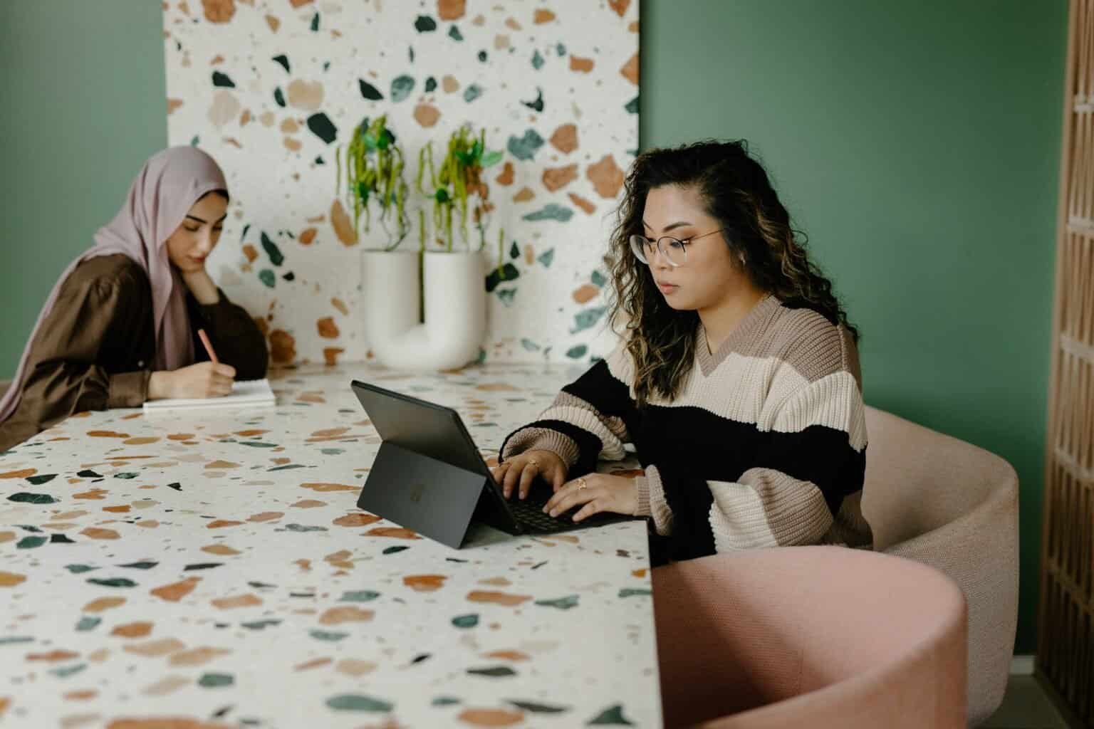 Two women working at a terrazzo table, one on a laptop, the other writing.