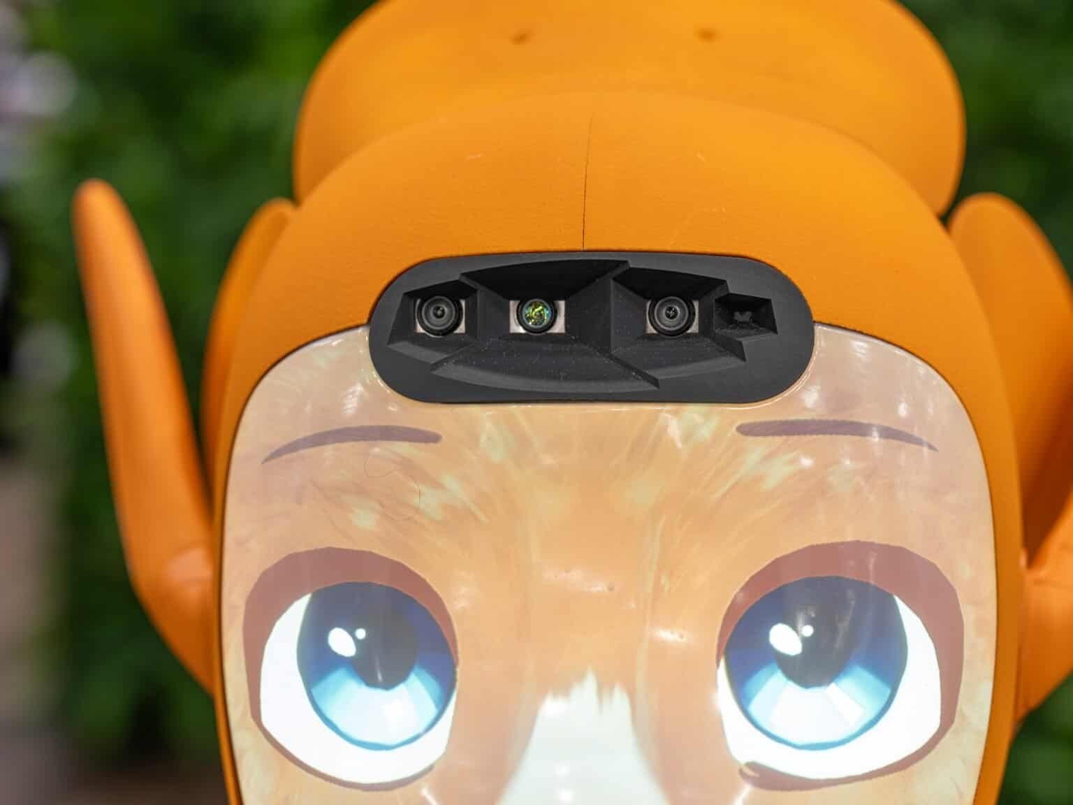 Close-up of a friendly orange robot's face with large blue eyes and a camera array.