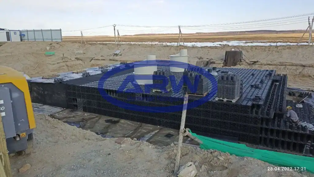 Crew assembling a large underground geocellular stormwater tank at a European jobsite—black crates with access risers on a lined and geotextile-protected excavation.
