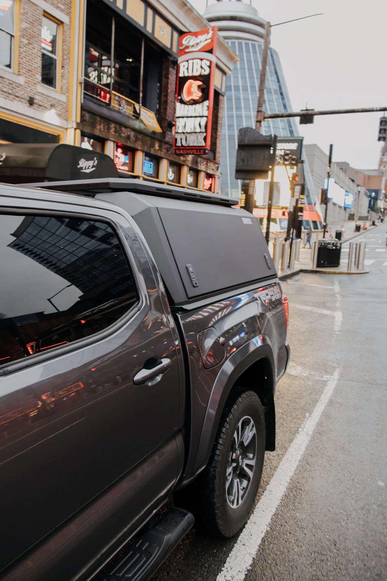 Stainless steel truck cap mounted on a dark gray pickup truck in an urban setting.