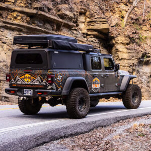 Stainless steel non-modular truck cap mounted on a rugged Jeep Gladiator parked alongside a rocky mountain road.