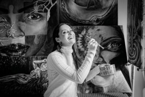 artist laughing while painting with paintings behind her. Black and white image.