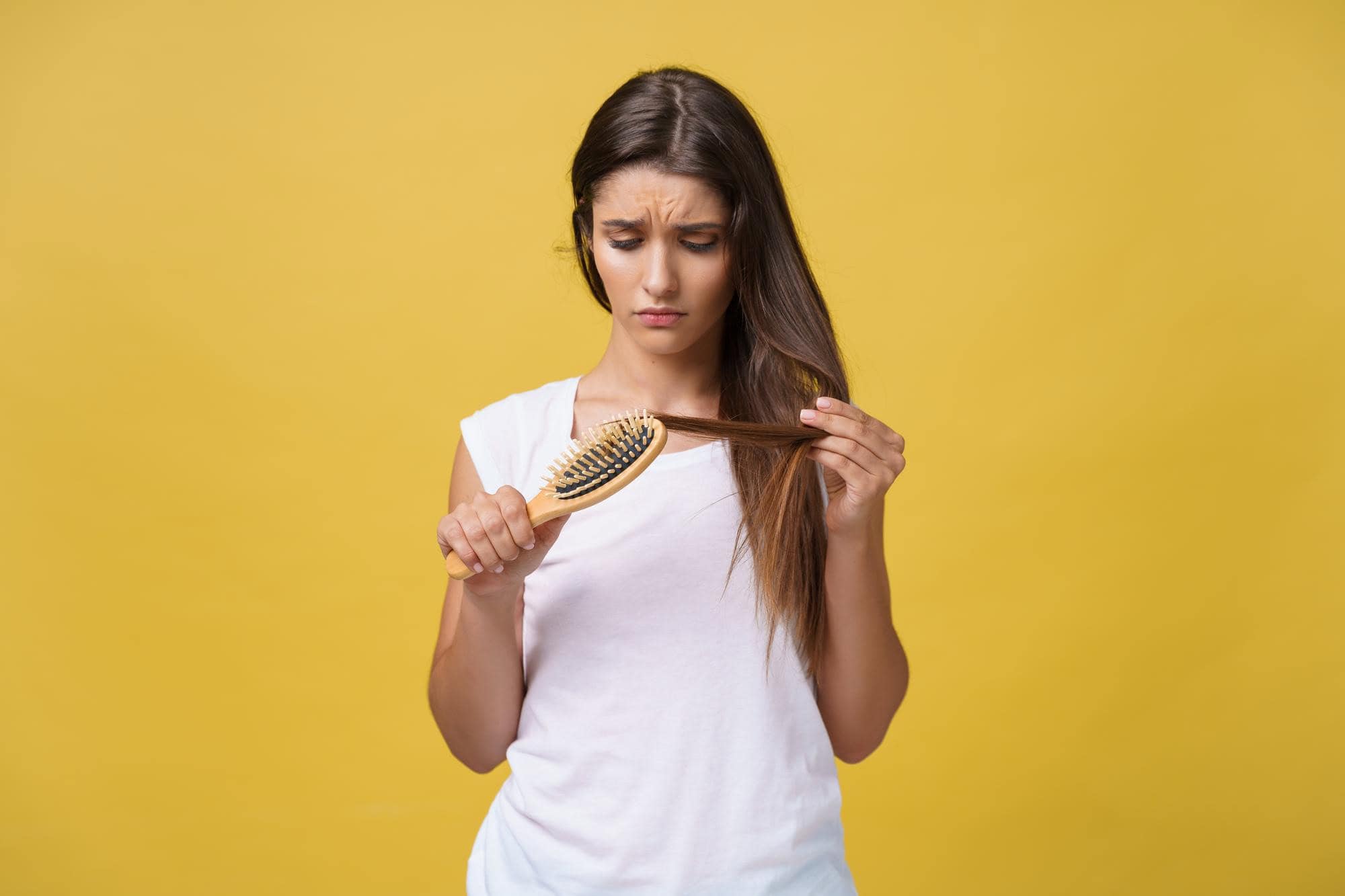 Discontent woman inspecting hair loss or damage while brushing hair against yellow background.