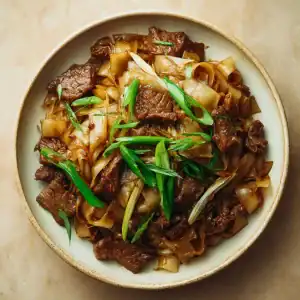 A bowl of Beef Chow Fun features stir-fried flat rice noodles with tender beef slices and green onions, served on a beige plate atop a light tan surface.
