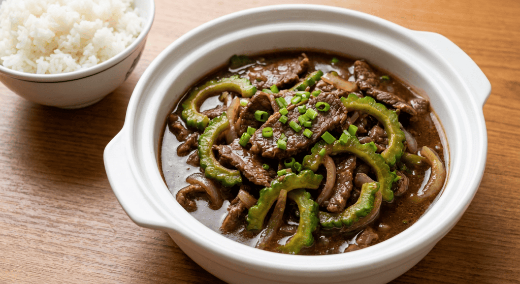 A bowl of Ampalaya Con Carne Recipe, featuring beef and bitter melon stir-fry in a dark sauce, garnished with chopped green onions, is served in a white dish next to steamed white rice on a wooden table.