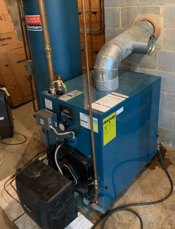 Blue boiler system in a basement with pipes and a vent. It is placed against a cinder block wall, surrounded by various equipment and boxes.