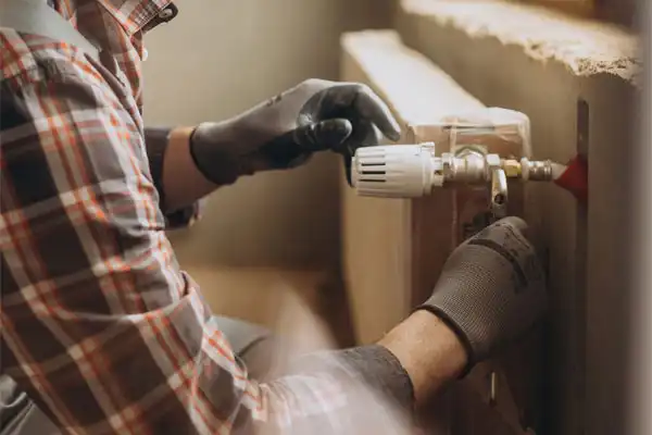Person wearing a plaid shirt and gloves adjusts a valve on a radiator in a room.