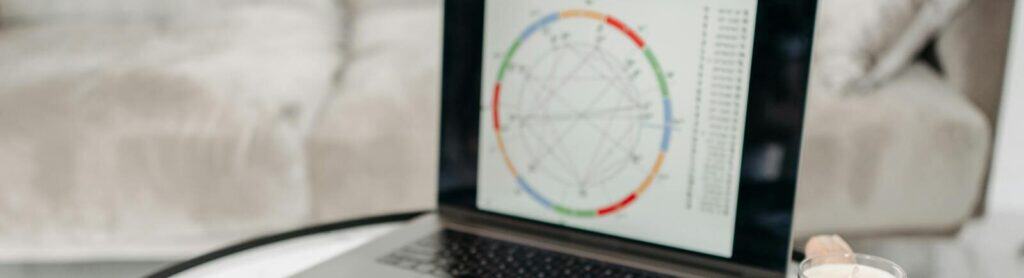 Astrology charts displayed on a laptop next to tarot cards and notebooks on a table.