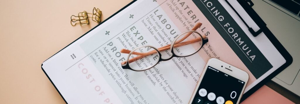 Overhead view of business tools including a phone calculator, pricing formula document, and eyeglasses on a desk.