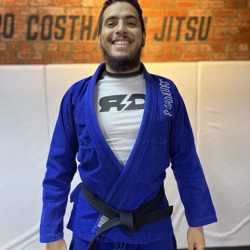 Head Coach Sharaf Brazilian Jiu-Jitsu black belt instructor in a blue gi smiling in front of a brick wall