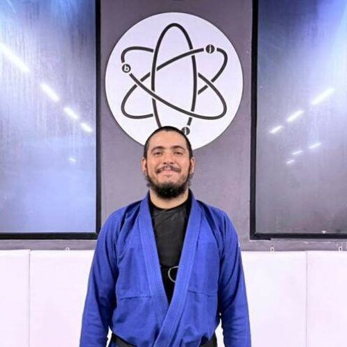 Atom Jiu Jitsu head coach standing in a blue gi in front of the Cicero Costha and Atom logos at the new academy in Cairo