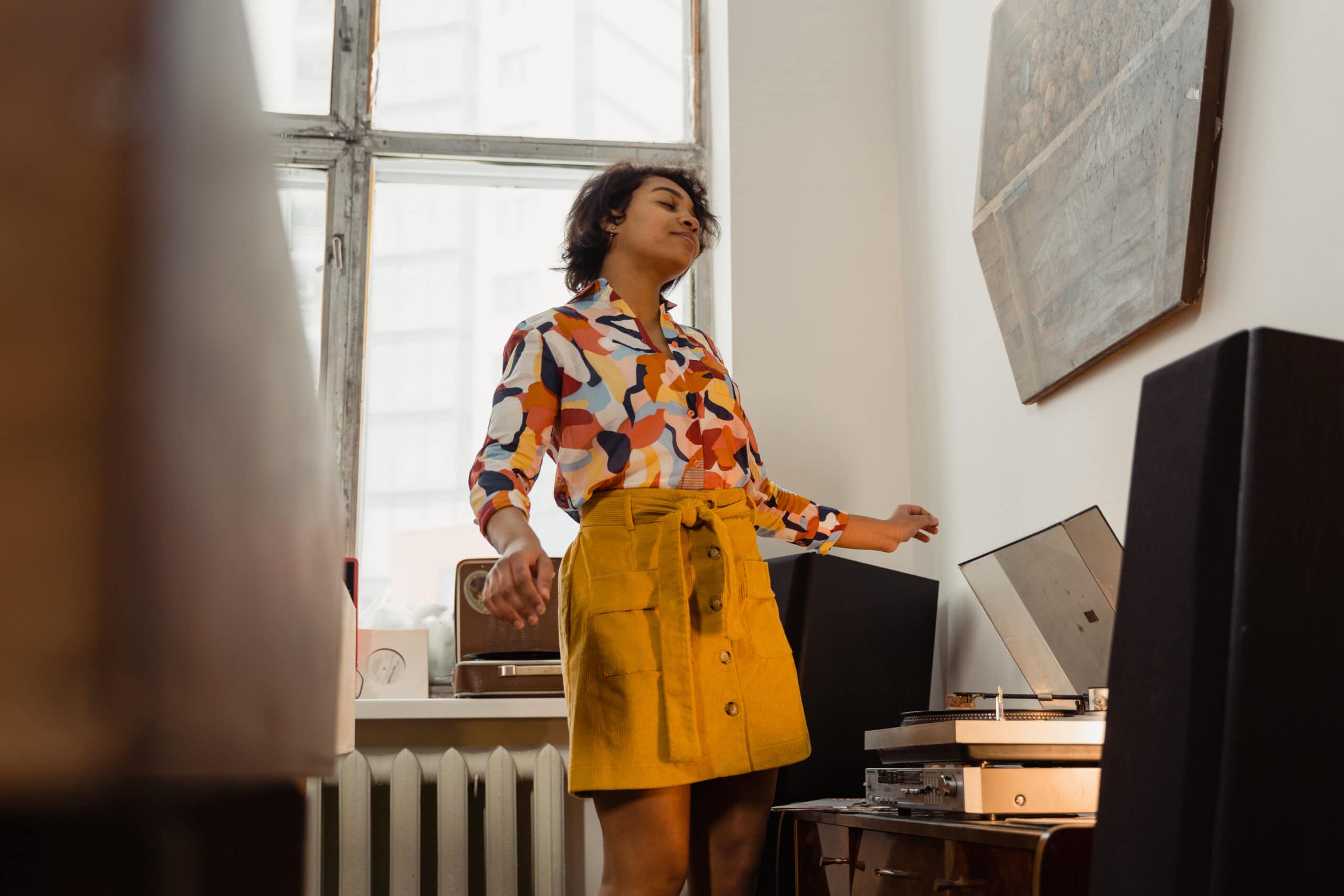 Using a premium home sound system, a young woman listens to music on a turntable with a smile on her face. She's wearing bright, sunny colors in front of a window.