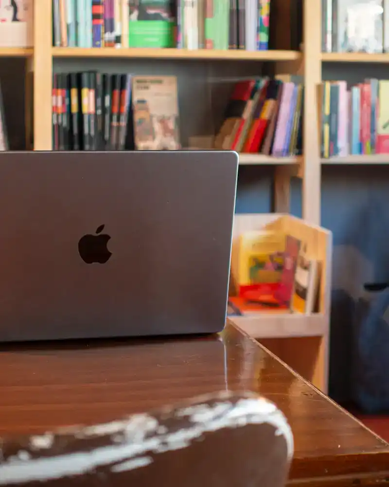 Apple MacBook partially open on a wooden table, with bookshelves filled with colorful books in the background.