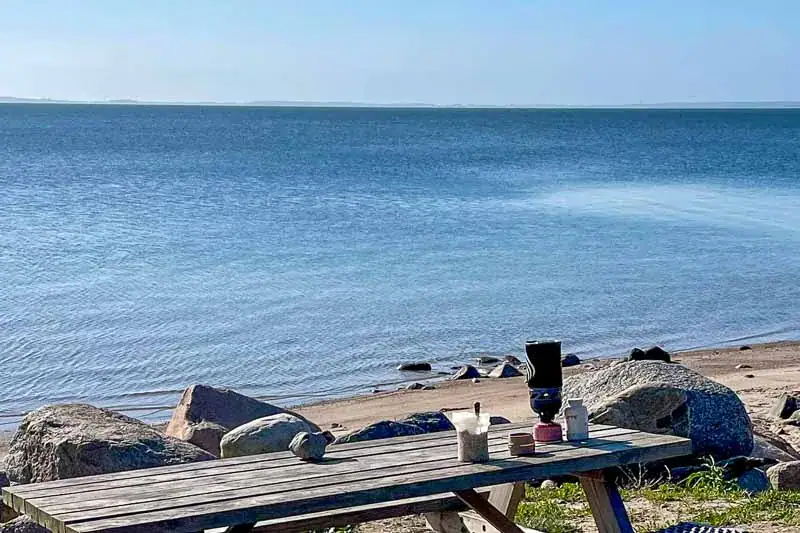 Bord ved stranden med udsigt over havet, med sten i forgrunden og en grøn markeret plads, hvor en brændstofkande og spisestel er synlige.