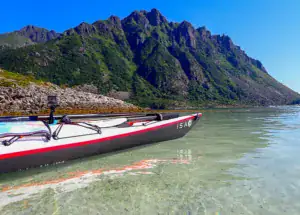 Havkajak ISA ligger ved stranden på lavt klart vand, klipper ses i baggrunden og himlen er klar blå