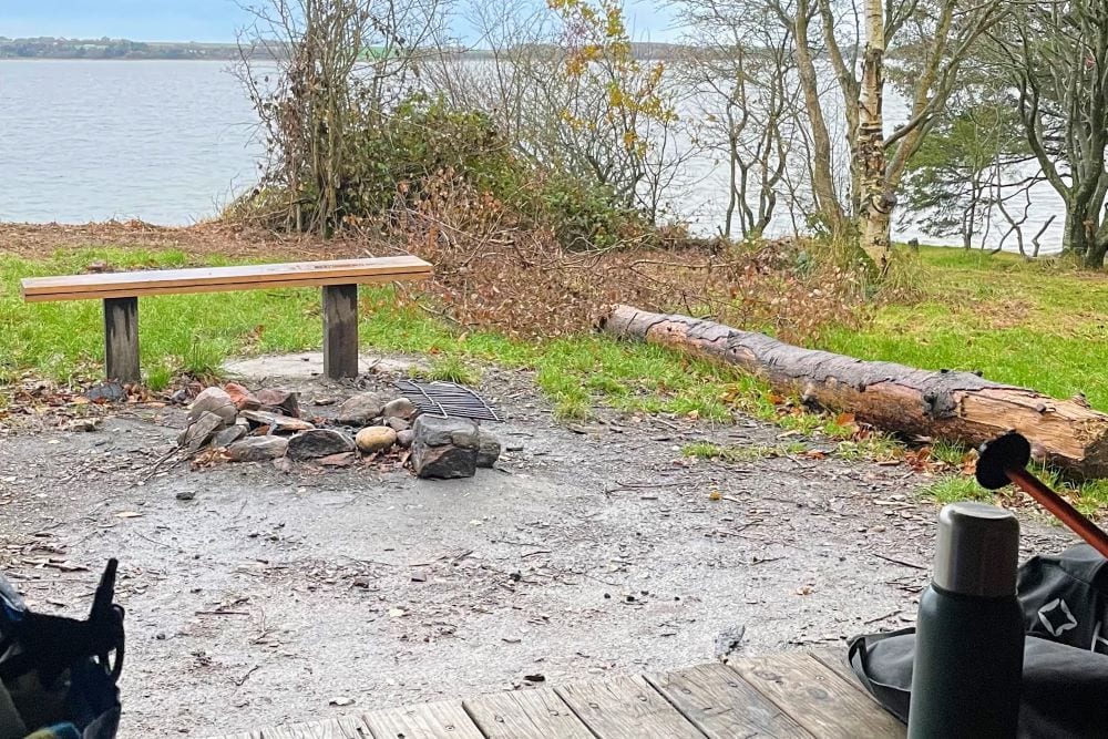 Billedet viser en rolig plads ved vandet med bålsted og siddeplads, perfekt til afslapning.