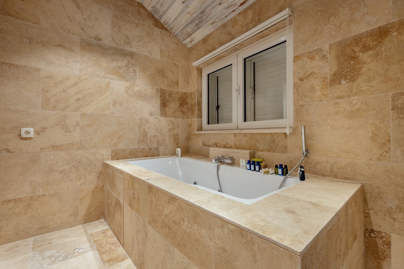 Luxury bathtub with beige stone walls and window in a modern bathroom setting.