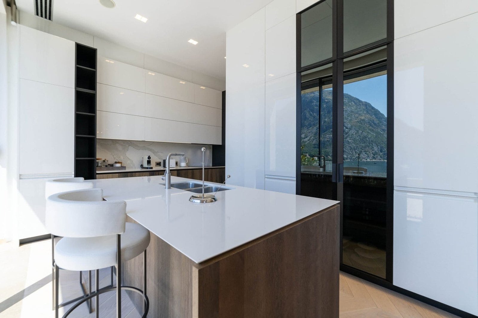 Modern minimalist kitchen with sleek white cabinets and mountain view through large glass window.