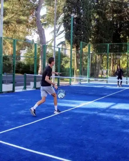 Tennissers die trainen op een blauwe buitenbaan, omringd door groene hekken en bomen.