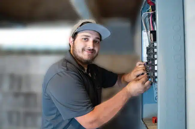 A skilled technician in a gray shirt and cap carefully services an electrical panel, ensuring safe, reliable comfort for your home.