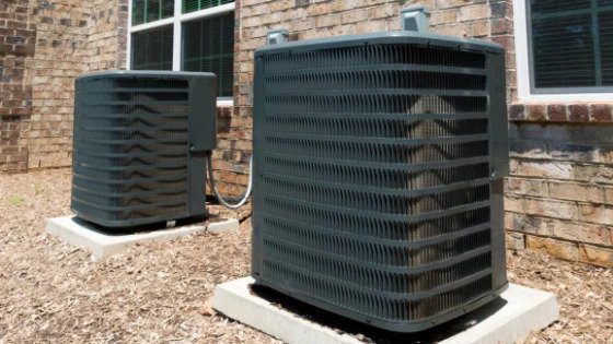 Two outdoor AC units on concrete pads by a brick wall, installed with expert care for 24/7 comfort and safety you can trust.