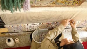 Woman is sitting in front of a loom and weaving Persian rug while she is looking at the rug map to choose the right colors.