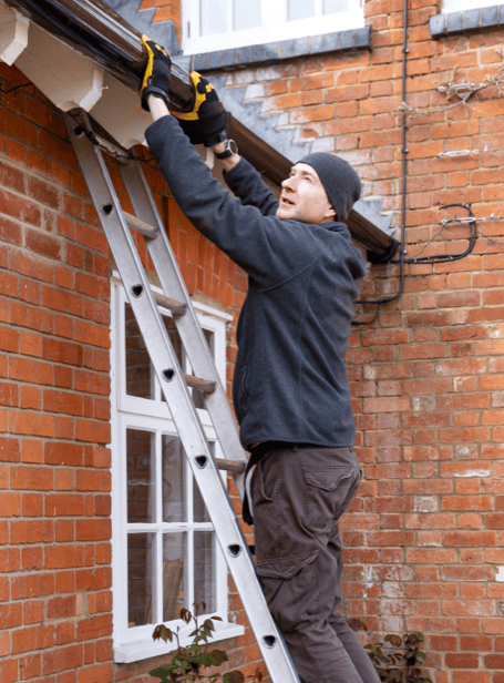 Man on ladder fixing gutter outside brick building at Blue Box Self Storage.