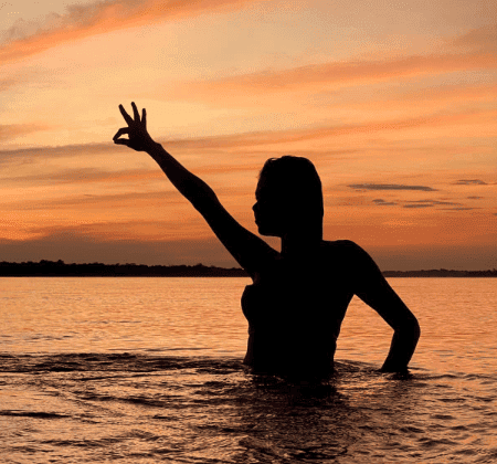 Woman in water at sunset with hand gesture, peaceful retreat scene.