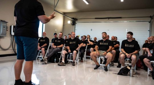 Men's Brotherhood gathering at Big Balls Brotherhood event, seated and listening attentively to a speaker in a garage-style meeting space.