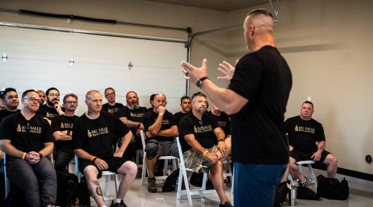 Men's group meeting at Big Balls Brotherhood event, attendees wearing matching black T-shirts with the organization's logo, listening to a speaker.