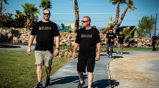 Bold text: Big Balls Brotherhood outdoor event with men wearing matching black t-shirts, walking in a scenic park with palm trees and rocks in the background.