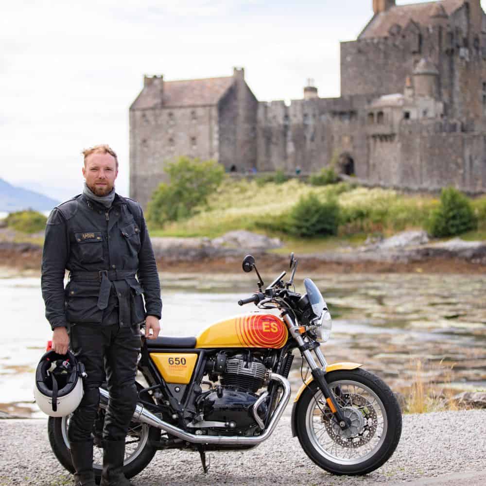 Motorcyclist wearing a wax cotton Belstaff jacket, with a Royal Enfield Interceptor 650, special Edition outside Eilean Donan Castle, on The West Coast of Scotland.