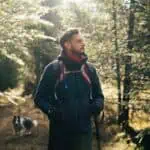 Ein Mann mit Rucksack und Trekkingstöcken geht durch einen sonnenbeschienenen Waldweg, und ein Hund folgt ihm.