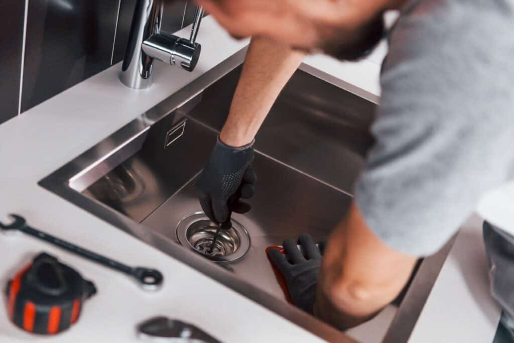 Plumber repairing kitchen sink drain with tools, emphasizing emergency plumbing services by Bizzy B Plumbing in Louisville.