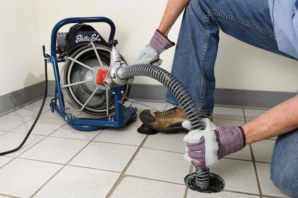 Plumber operating Electric Eel drain cleaning equipment, addressing clogged drain in tiled floor setting.