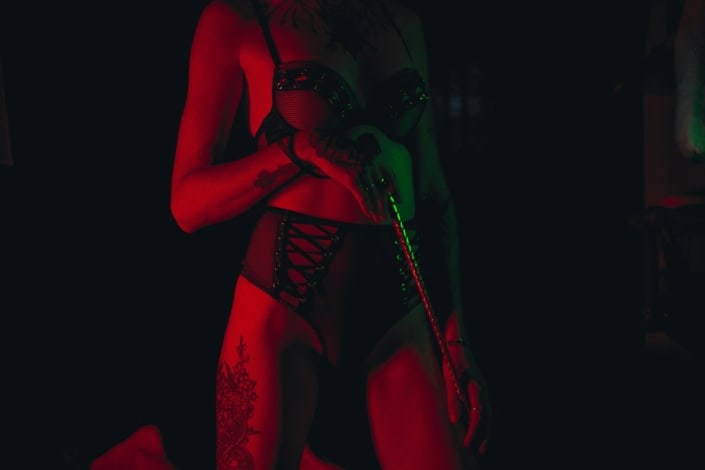 Mulher segurando um chicote, em um cenário hot Mulher em pé segurando um chicote em um cenário de sadismo