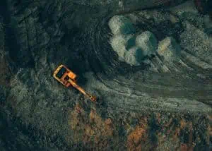 Aerial view of a mining site featuring a large orange excavator working on the ground. The excavator is positioned on a dark, earthy surface with visible tire tracks and excavation marks. Adjacent to the excavator are several mounds of mined material, showcasing the ongoing excavation process. The landscape around the site is rugged, with various shades of brown and gray, indicating different mineral compositions and geological features. This image visually represents the mining industry's operations and the importance of precise analytical techniques in assessing and processing mineral compositions.