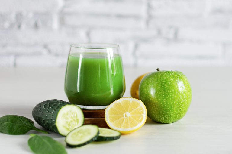 Fresh green cucumber, lemon, and apple juice in a glass on a white wooden surface with ingredients around it.