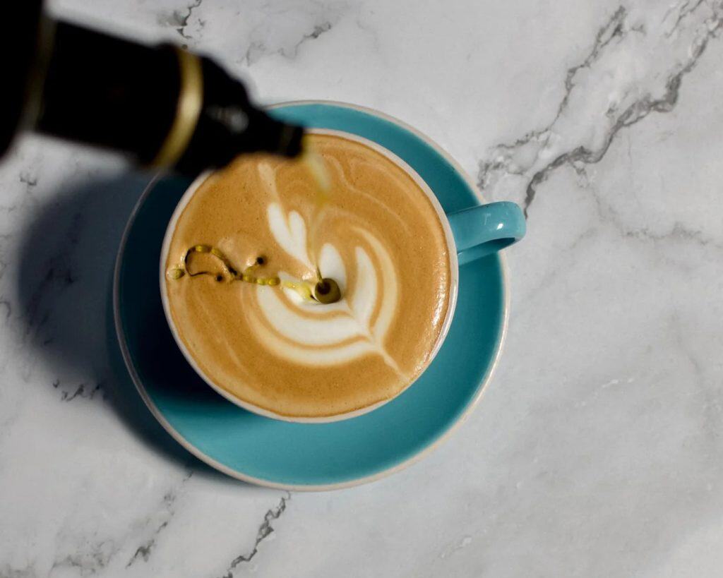 Creamy coffee latte with heart-shaped foam art being drizzled with honey in a blue mug on a marble surface.