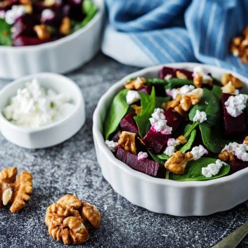 Fresh beet and spinach salad with walnuts and goat cheese in white ceramic bowls, served with additional toppings on rustic surface.