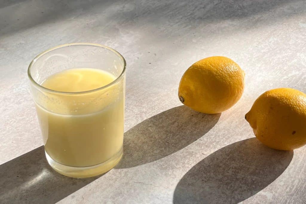 Freshly squeezed lemon juice in a glass with two whole lemons on a light-colored surface under natural sunlight.