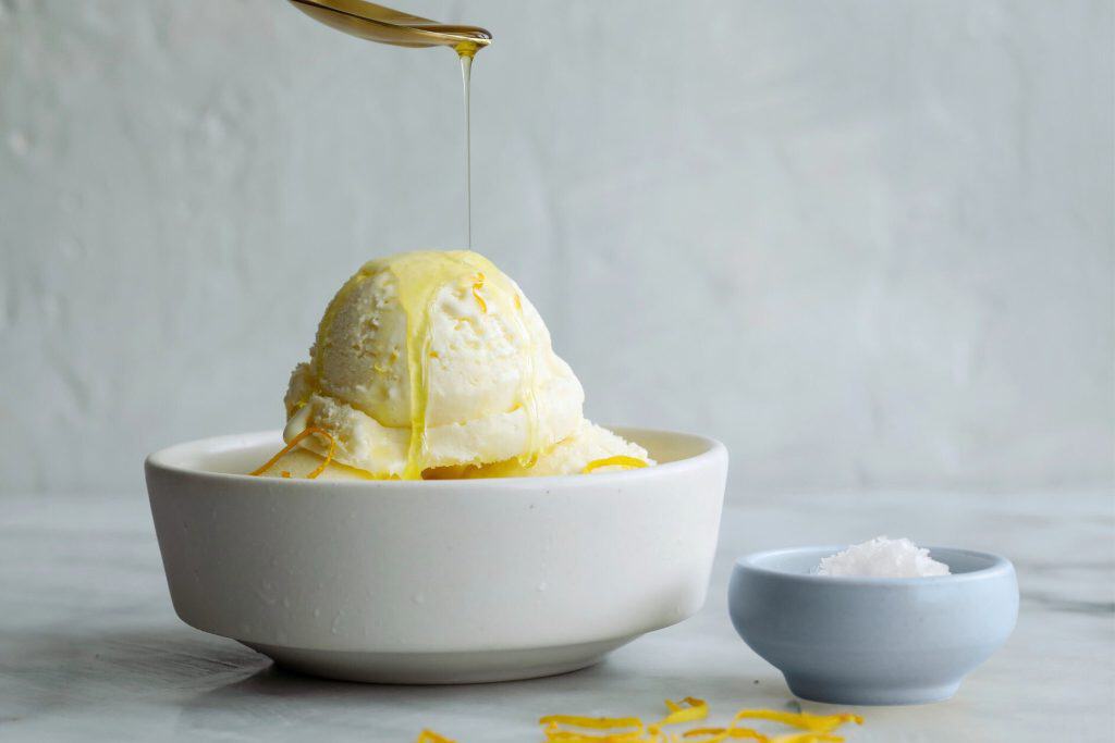 Creamy vanilla ice cream with lemon zest drizzle in a white bowl, served on a marble surface with a small salt bowl nearby, creating a refreshing summer dessert.