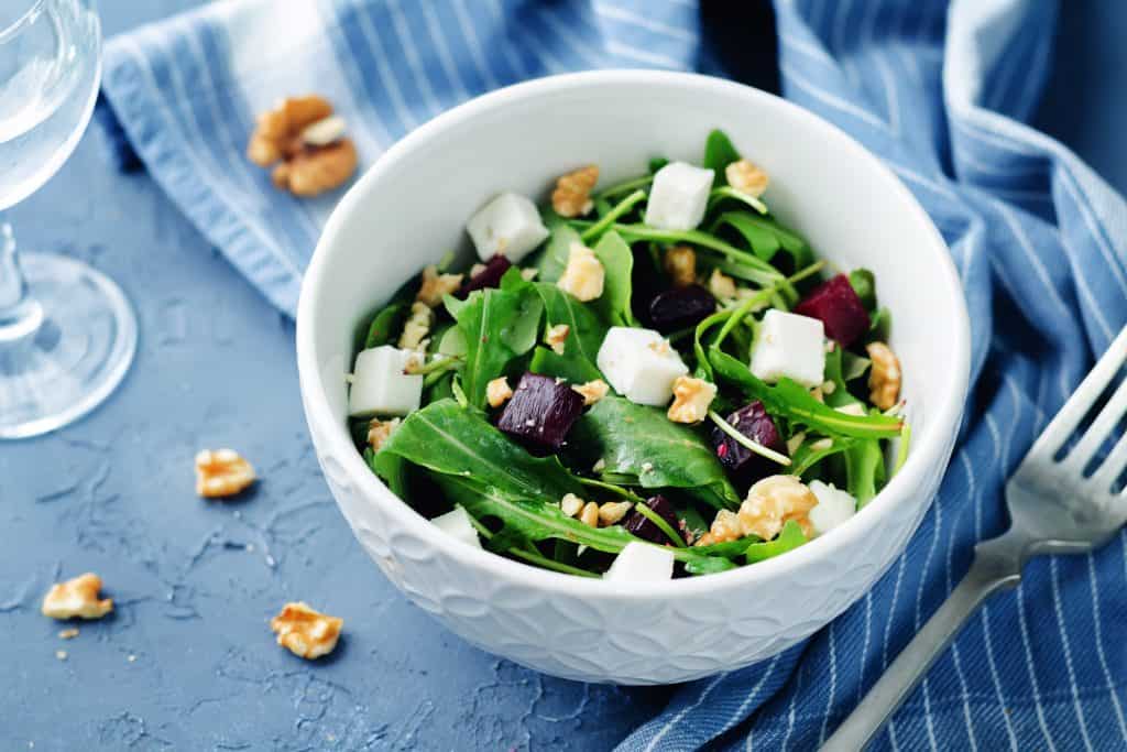 Fresh spinach salad with feta cheese, walnuts, and beets in a white bowl on blue textured surface.