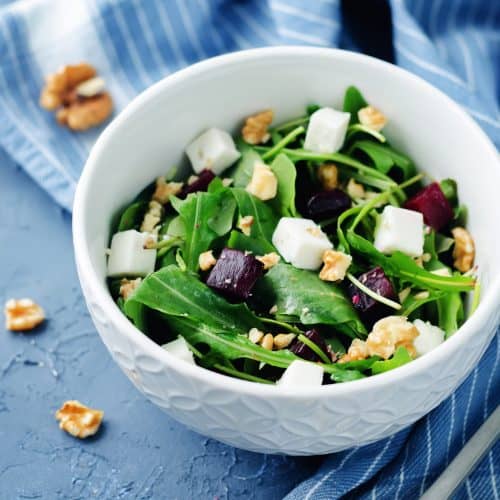 Fresh spinach salad with feta cheese, walnuts, and beets in a white bowl on blue textured surface.