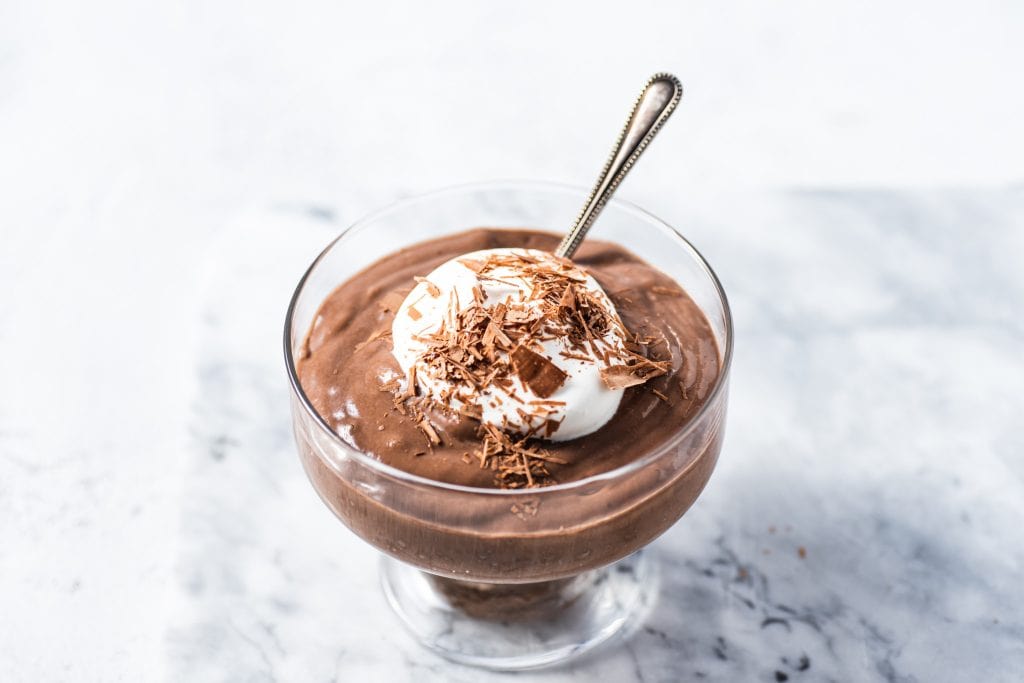 Rich chocolate mousse topped with whipped cream and chocolate shavings in a glass dessert cup on a white marble surface. Perfect for chocolate lovers and dessert enthusiasts.